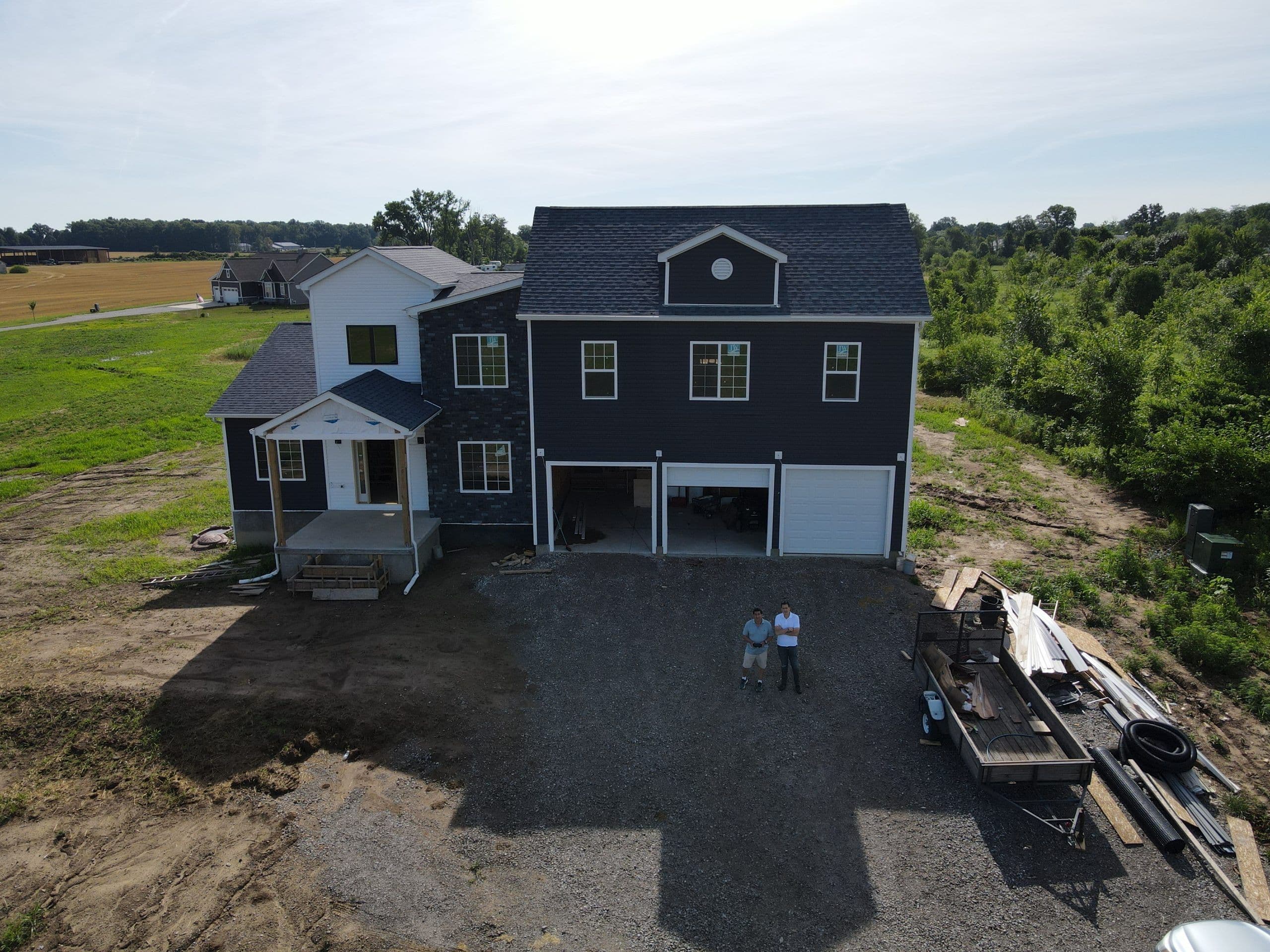 Aerial view of a Central Ohio home partway through construction, charcoal siding and a three-car garage wing, with two of the crew standing on the gravel drive.